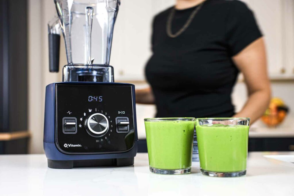 Woman walking behind a Vitamix blender next to two green smoothies.